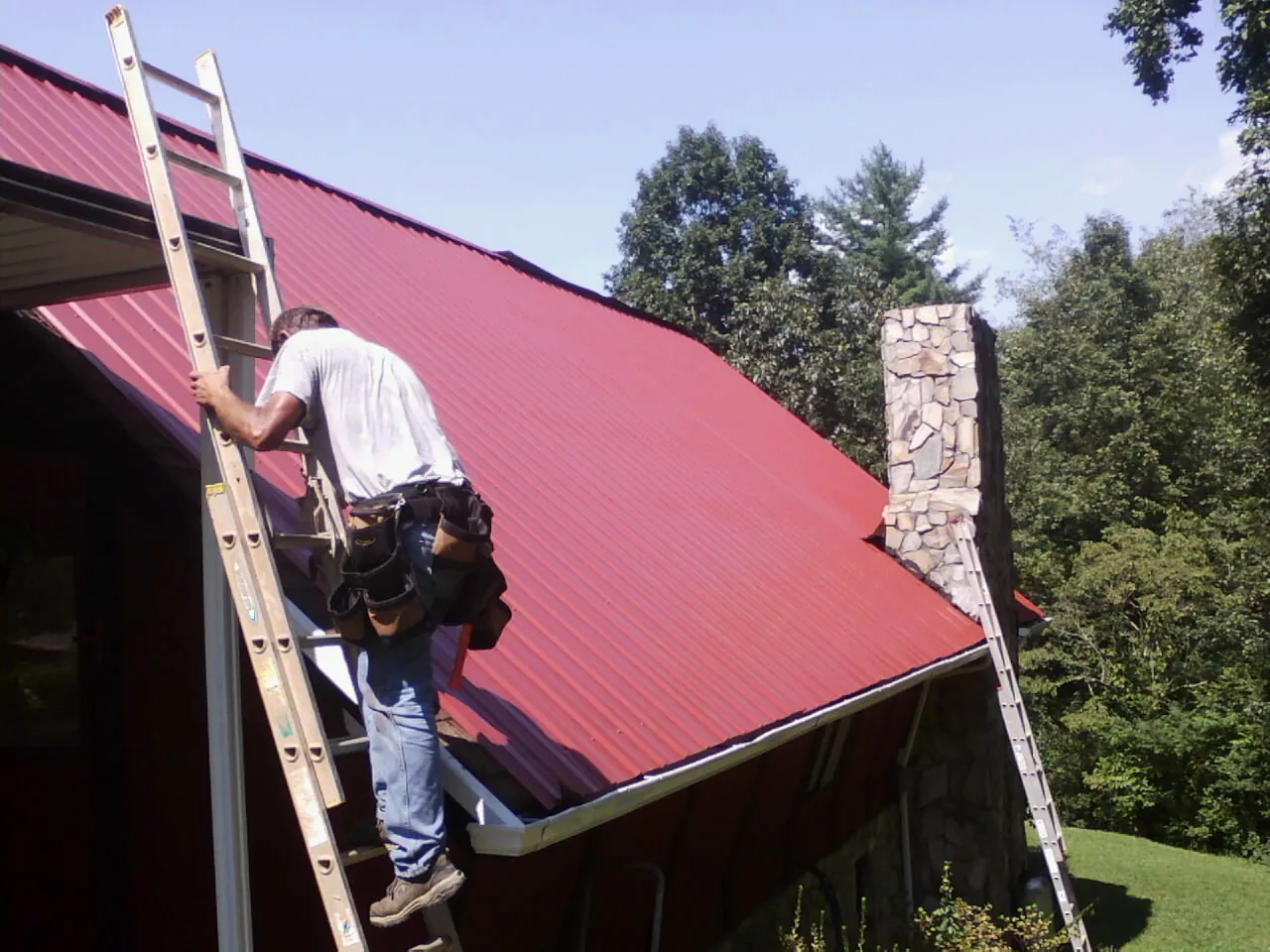 Roofer on a ladder beside a red metal roof and stone chimney — Soffit & Fascia Repair by Roofing Brothers in Peotone